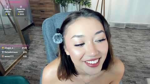 A young Asian woman with shoulder-length brown hair, wearing a silver hair clip, sits on a chair, smiling, with a video overlay showing chat messages.
