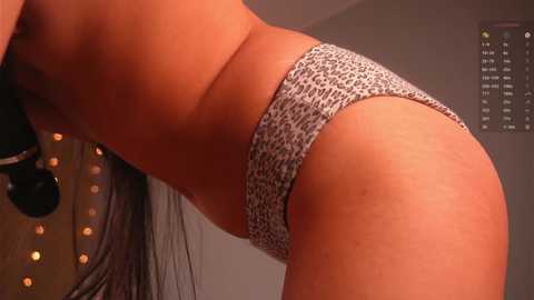 A close-up video of a woman with medium skin tone wearing leopard print panties, leaning forward with long, straight black hair, in a dimly lit room with string lights and a clock on the wall.