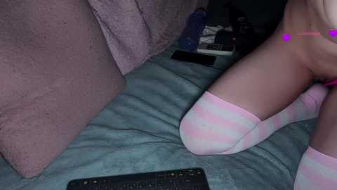 Video of a light-skinned woman in pastel striped socks kneeling on a teal blanket, wearing a pink necklace. Background includes a purple couch, a laptop, and a TV remote.