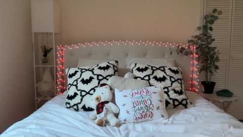 Video of a cozy bedroom with a white tufted headboard, white bedding, a teddy bear, and two decorative pillows with black bat and red string light borders.