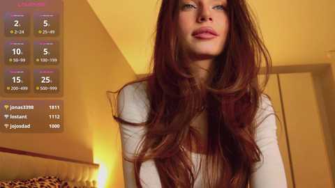 Video of a Caucasian woman with long, wavy auburn hair, light skin, and full lips, wearing a white top, standing in a warmly-lit bedroom.