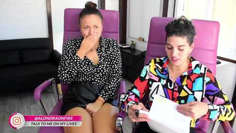 Video: Two women in modern chairs, one wearing a black patterned top and the other a colorful abstract top, discuss a document.