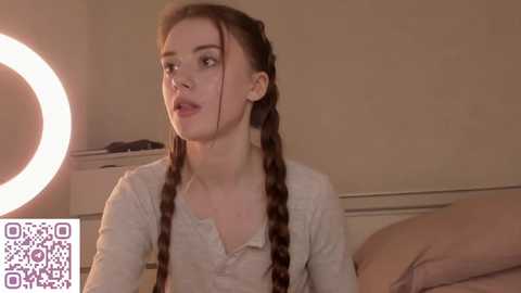 A video of a young woman with long brown braids, pale skin, and wearing a light-colored top, seated in a softly lit room with a round light on the left and a bed behind her.