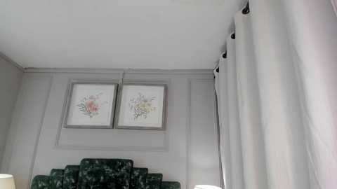 Video of a minimalist bedroom with white walls, two floral prints, and a green velvet headboard. White curtains with black tassels hang from the right side.