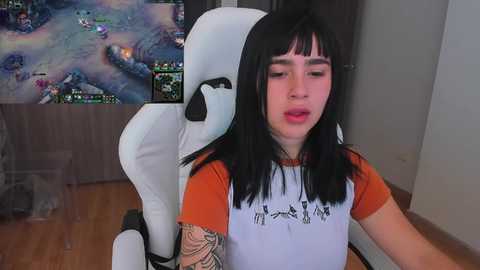 Video of a young woman with long black hair, wearing an orange and white shirt, playing League of Legends on a white gaming chair. Background includes a TV displaying the game and a wooden floor.