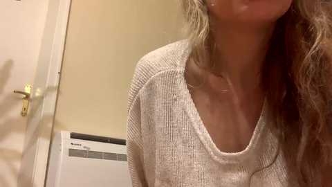 Video of a young woman with long, wavy blonde hair, wearing a loose-fitting, beige sweater, standing in a beige-walled room with a white door and a vent.