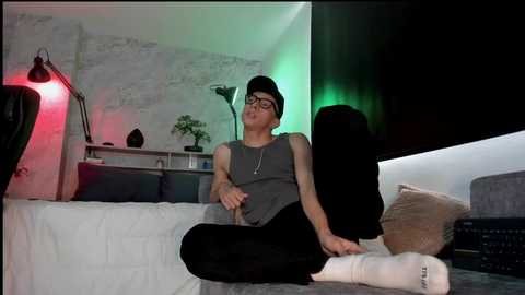 Video of a slender Asian man with glasses and a black hat, sitting on a sofa in a dimly lit room with green and red lighting, wearing a gray tank top and black pants.