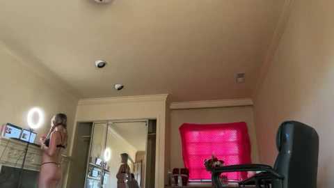 Video of a modern, beige-painted room with a large mirror, pink inflatable, and a black chair. A woman with light brown hair, wearing a beige bikini, stands near a vanity mirror.