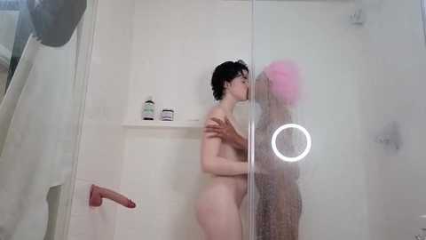Video of a nude woman with short black hair and a pink wig kissing through a shower glass. She has a small bust, fair skin, and is in a white-tiled shower with a ring light.