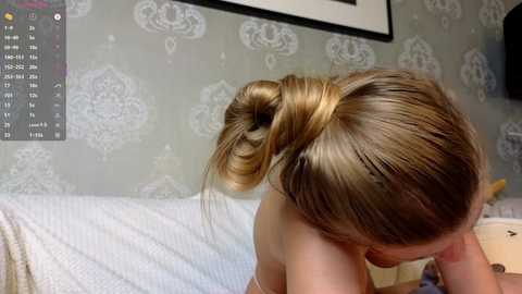 A video of a blonde woman with a bun hairstyle, topless, on a bed with white and grey floral-patterned wallpaper in the background.