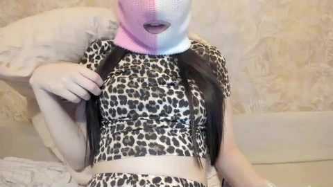 Video of a person wearing a leopard print crop top, pink and blue face mask, and black leather jacket. Background shows a beige, patterned wall and white bed with a pillow.