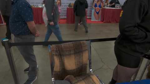 Video of a crowded indoor event with a person reclining in a chair, watching a performer on stage; people in casual attire, red drapes, and a stage backdrop.