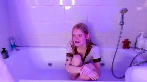 Video of a smiling young woman with blonde hair, wearing a white shirt, sitting in a bathtub under purple lighting.