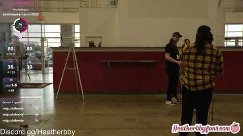 Video of a gym with a man in a yellow plaid shirt facing a woman in a black top. Virtual workout display on the left.