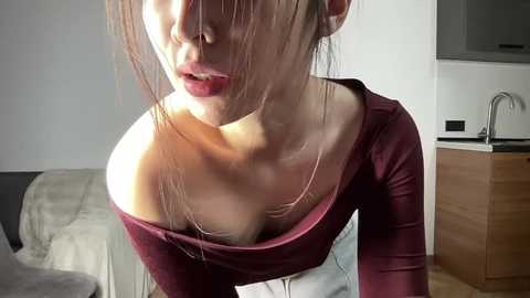 Video of an Asian woman with fair skin, red lipstick, and brown hair, wearing a deep burgundy off-shoulder top, leaning forward in a modern kitchen with white cabinets and a stainless steel sink.