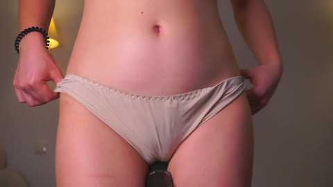 Video of a woman's torso, focusing on her beige panties, held slightly down by her hands, with a blurred indoor background.