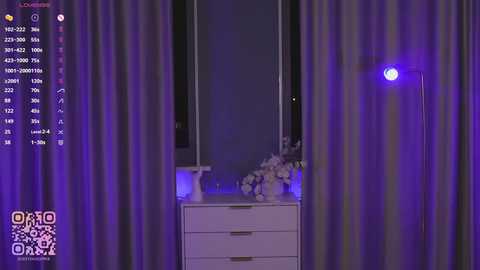 A video of a modern bedroom with purple curtains, a white dresser, a small mirror, and a glowing blue lamp on the nightstand.
