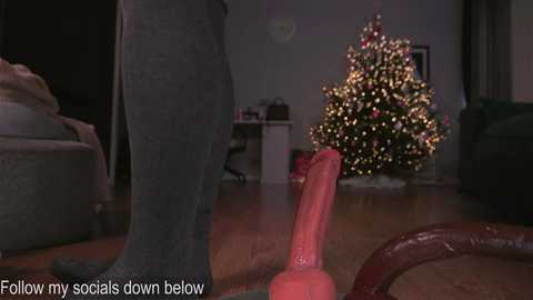 Media: Video of a Christmas tree lit with white lights, a red dildo on the floor, and a person's leg in gray socks, set in a dimly lit living room. Text at the bottom reads: \"Follow my socials down below.\