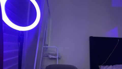 A video of a dimly lit room with a large, glowing white circular light on the left wall, casting a blue hue. A small white chair and a black-framed mirror with a white cord hang on the right side.