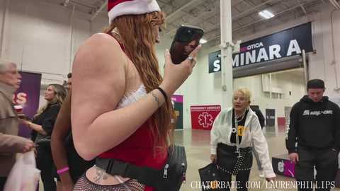 Video of a plus-size woman in a red Santa hat, black shirt, and fishnet tights, walking with a phone, in a busy airport, surrounded by diverse travelers.