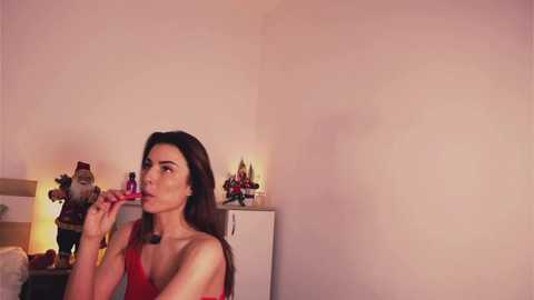 Video of a fair-skinned woman with long brown hair, wearing a red dress, applying lipstick in a softly lit, minimalist bedroom with white walls and a cluttered nightstand.