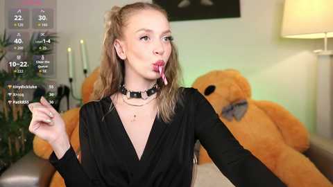 Video of a young woman with blonde hair, wearing a black dress and black choker, licking a lollipop, in a modern room with green walls, a lamp, and a large teddy bear.
