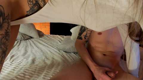 Video of a tattooed woman, partially nude, lying on a bed with striped sheets. Her left arm and torso are visible, displaying a small breast and intricate tattoos. The background features a brick wall and a white canopy.