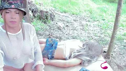 Video of a young woman in a white mesh top and blue shorts, lying face-down on a picnic table, with a man in a cap and t-shirt standing nearby in a grassy, forested area.