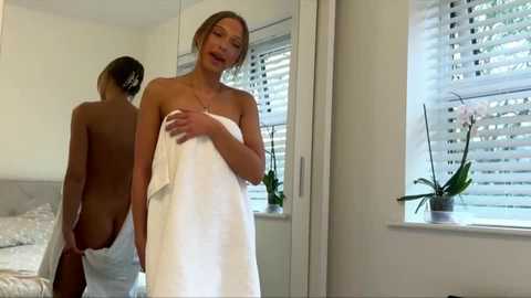 A video of a slender, topless woman with light skin and medium-length blonde hair, wrapped in a white towel, standing in a bright, modern bedroom with white walls, a bed, and a window with blinds.