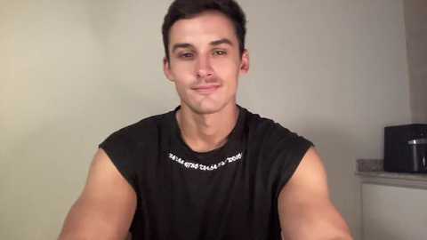 Video of a young, muscular man with short dark hair, wearing a black sleeveless shirt with white text, smiling, in a simple indoor setting with neutral-colored walls and a black object on the right.