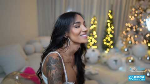 Video of a smiling Latina woman with long black hair, wearing a white lace bralette, adorned with tattoos, standing in a festive living room with a Christmas tree and decorations.