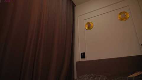 A video of a beige wall with a closed door, featuring two round, yellow light switches on the left and a black electrical outlet on the right. The background shows a dark, textured curtain.