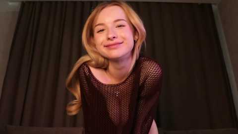 Video of a smiling, fair-skinned, blonde woman wearing a black fishnet top, standing in front of dark curtains.