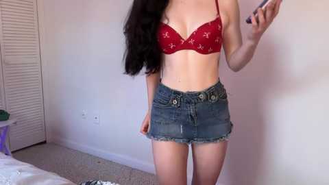 Video of a slender, fair-skinned woman with long black hair, wearing a red lace bra and frayed denim skirt, taking a selfie in a simple bedroom with white walls and a carpeted floor.
