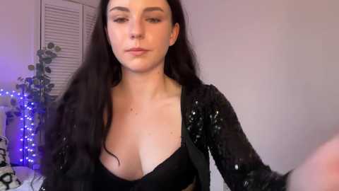 Video of a light-skinned woman with long, wavy black hair, wearing a black sequined top that reveals ample cleavage, standing indoors with a plant and blue string lights in the background.