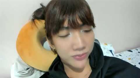 Video of a young Asian man with short brown hair, wearing a black hoodie, lying on a bed with a plush donut pillow, eyes closed, smiling softly.