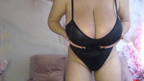 Video of a voluptuous, light-skinned woman in a black, high-cut swimsuit, showcasing large breasts. Background features a light-colored wall and a pink floral-patterned fabric.
