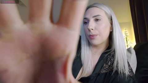 Video of a fair-skinned woman with long platinum hair, wearing a black lace top, gazing at a large hand in the foreground, indoors.