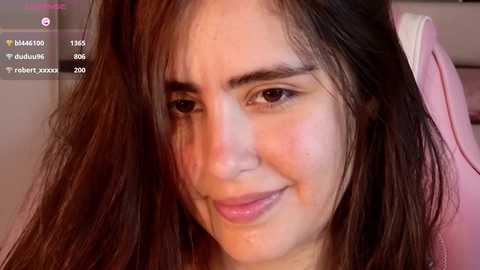 Video of a young woman with long, dark hair and fair skin, wearing pink clothing, smiling slightly with a slight blush on her cheeks. The background is blurred, suggesting an indoor setting.