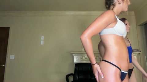 Video of two women in bikinis, one in a white sports bra and black thong, the other in a blue top and black thong, in a beige-walled room with a black office chair and white electrical outlet.