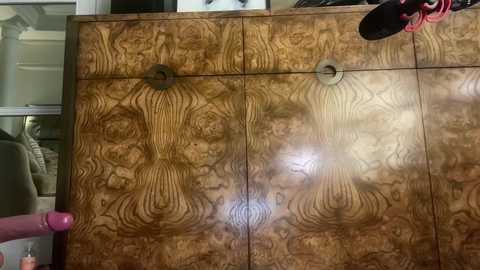 Video of a large, ornate wooden wardrobe with intricate grain patterns and three circular handles, partially open to reveal a pink garment. Background includes a beige chair and partially visible white walls.