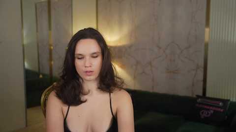 Video of a fair-skinned woman with long, dark hair, wearing a black spaghetti-strap top, seated in a dimly-lit, elegant room with marble walls and a green couch.