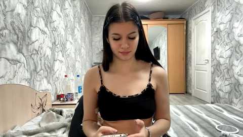 Video of a young woman with long black hair, wearing a black camisole, sitting on a bed in a room with grey and white marble-patterned wallpaper, wooden furniture, and a door.