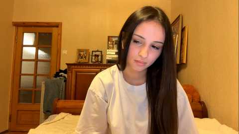 Video of a young woman with long dark hair, wearing a white shirt, sitting on a bed in a warm, beige room with wooden furniture and framed pictures.