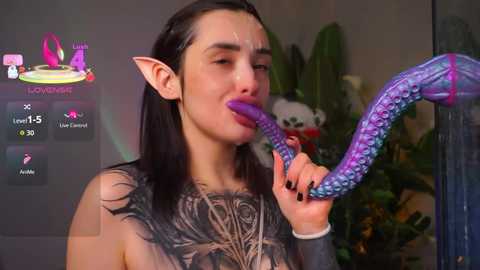A video of a young woman with long black hair and pointed elf ears, wearing a black top with a detailed tattoo, licking a purple, tentacle-like object. Background includes a large potted plant and a blurred room.