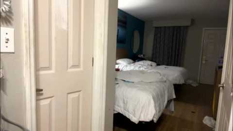A video of a dimly-lit, cluttered hotel room with two unmade beds, a blue wall, and closed white door, featuring a messy floor.