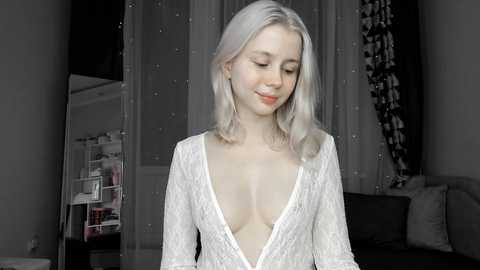 A video of a young, fair-skinned, blonde woman with shoulder-length hair, wearing a low-cut, white lace dress, standing in a dimly lit room with a mirror and shelves in the background.