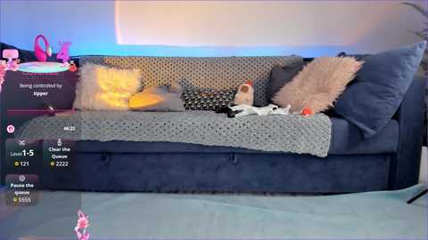 Video of a plush, dark blue couch with a gray knitted throw, adorned with cushions and a stuffed toy. Background includes a pink bag and blue lighting, suggesting a cozy, lived-in room.