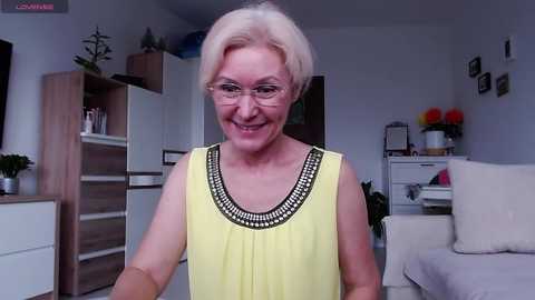 Video of an elderly Caucasian woman with short, platinum blonde hair, wearing glasses and a yellow blouse with black trim, smiling in a modern living room with white furniture and plants.