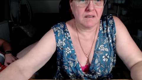 Video of a middle-aged, fair-skinned woman with glasses and a floral-patterned blue dress, seated in a dimly lit room, holding a red object, possibly a phone.